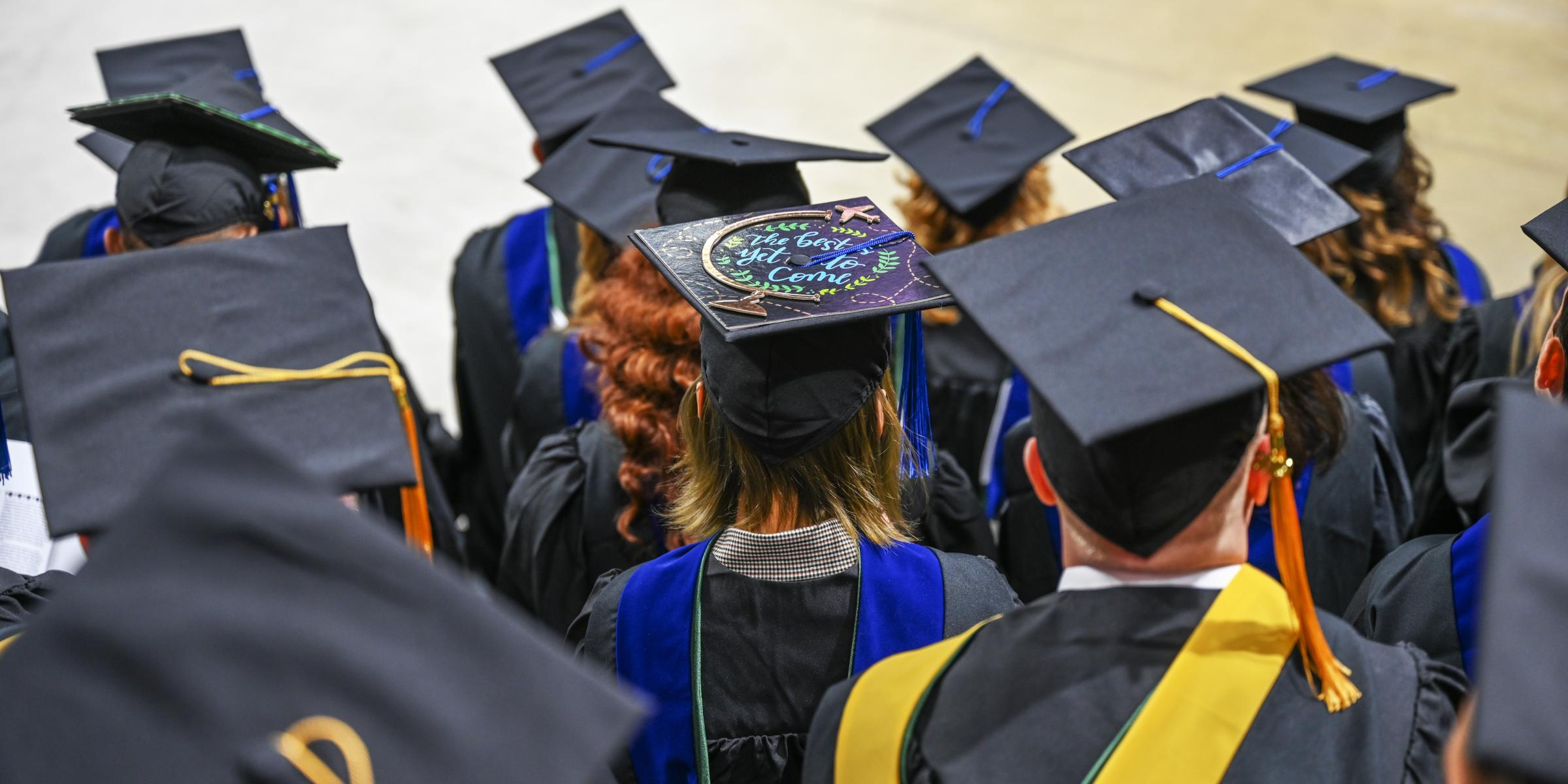 A group of graduating students in cap and gowns facing away from the camera. The center student's cap reads "The best is yet to come."